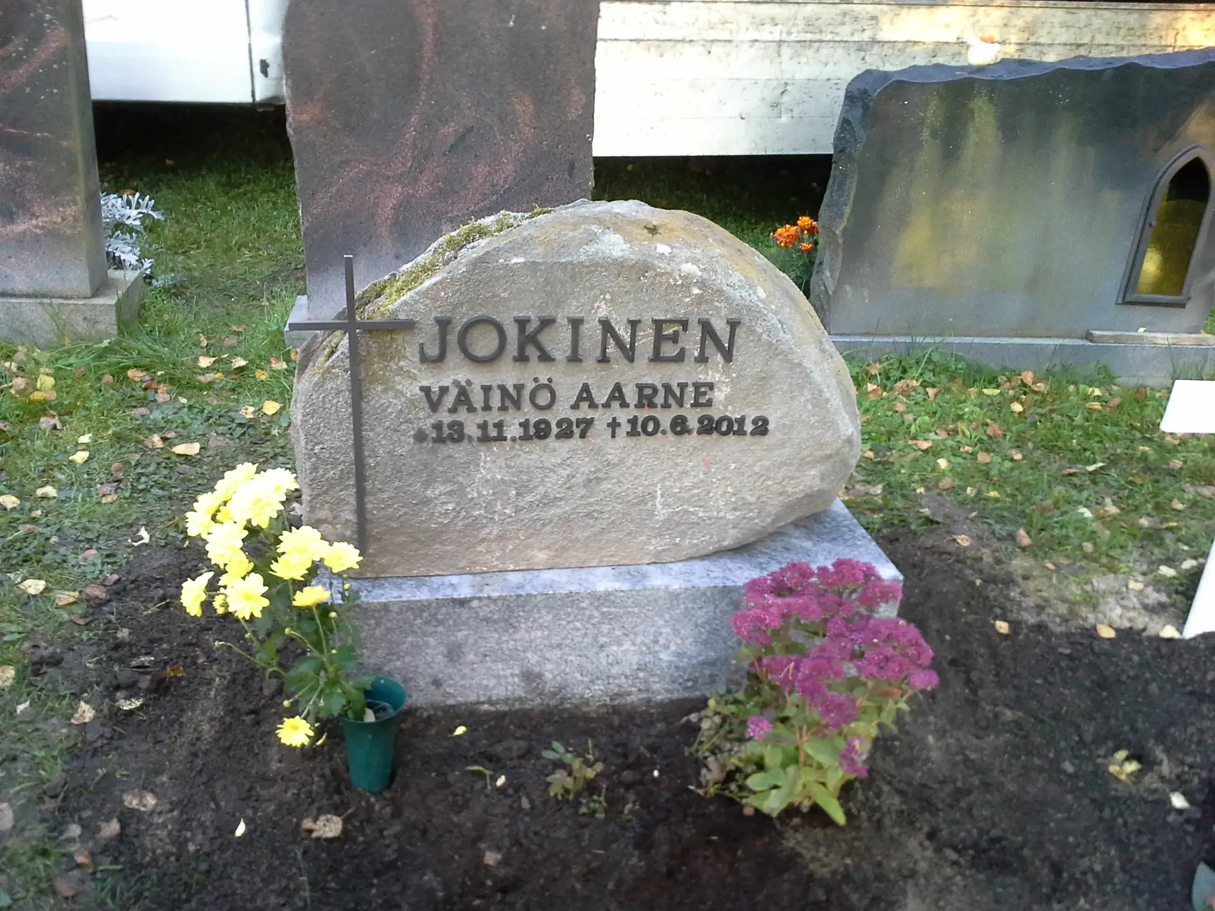 Lähikuva hautausmaan hautakivestä. Keskellä on suuri, vaaleanharmaa, sammalpeitteinen luonnonkivi, jossa on tummat metallikirjaimet "JOKINEN VÄINÖ AARNE 13.11.1927 +10.6.2012". Kiven vasemmalla puolella on tumma metalliristi. Kivi seisoo vaaleanharmaalla graniittijalustalla. Kiven edessä vasemmalla on ryhmä kirkkaankeltaisia kukkia ja oikealla purppuranpunaisia maksaruohoja. Taustalla näkyy vihreää ruohoa, ruskeita syyslehtiä ja muita hautakiviä, sekä osittain valkoinen rakennus. Kuva on otettu luonnollisessa päivänvalossa.