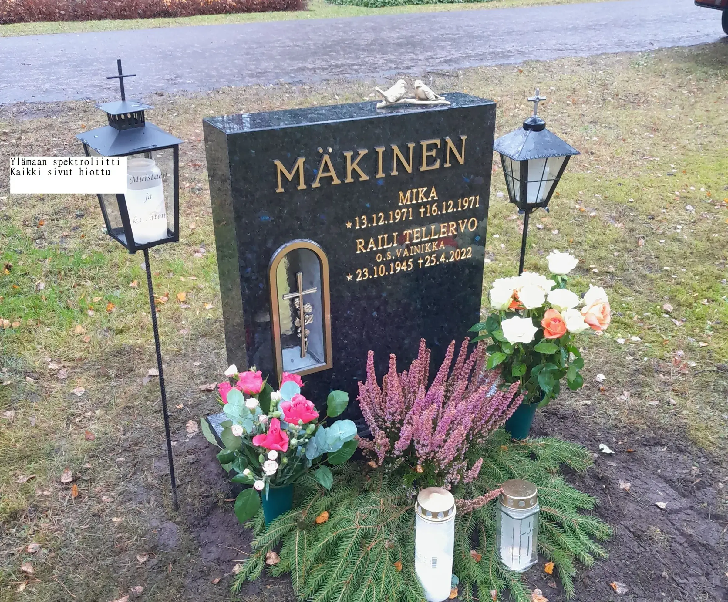 Tumma graniittinen hautakivi kultaisilla kirjaimilla, joissa lukee "MÄKINEN", "MIKA *13.12.1971 +16.12.1971" ja "RAILI TELLERVO O.S. VAINIKKA *23.10.1945 +25.4.2022". Kiven päällä on kaksi linnunhahmoa ja keskellä syvennys, jossa on risti ja ruusu. Hautakiven molemmin puolin on mustat lyhdyt, joissa on ristit yläosassa. Vasemmassa lyhdyssä palaa kynttilä, jossa on teksti "Muistaen ja kaivaten". Hautakiven edustalla on vaaleanpunaisia ruusuja, violettia kanervaa ja havunoksia, joilla on kaksi lasista hautakynttilää. Oikealla on valkoisia ja oransseja ruusuja. Maassa on nurmikkoa ja pudonneita lehtiä, taustalla märkä asfalttitie pilvisenä päivänä.
