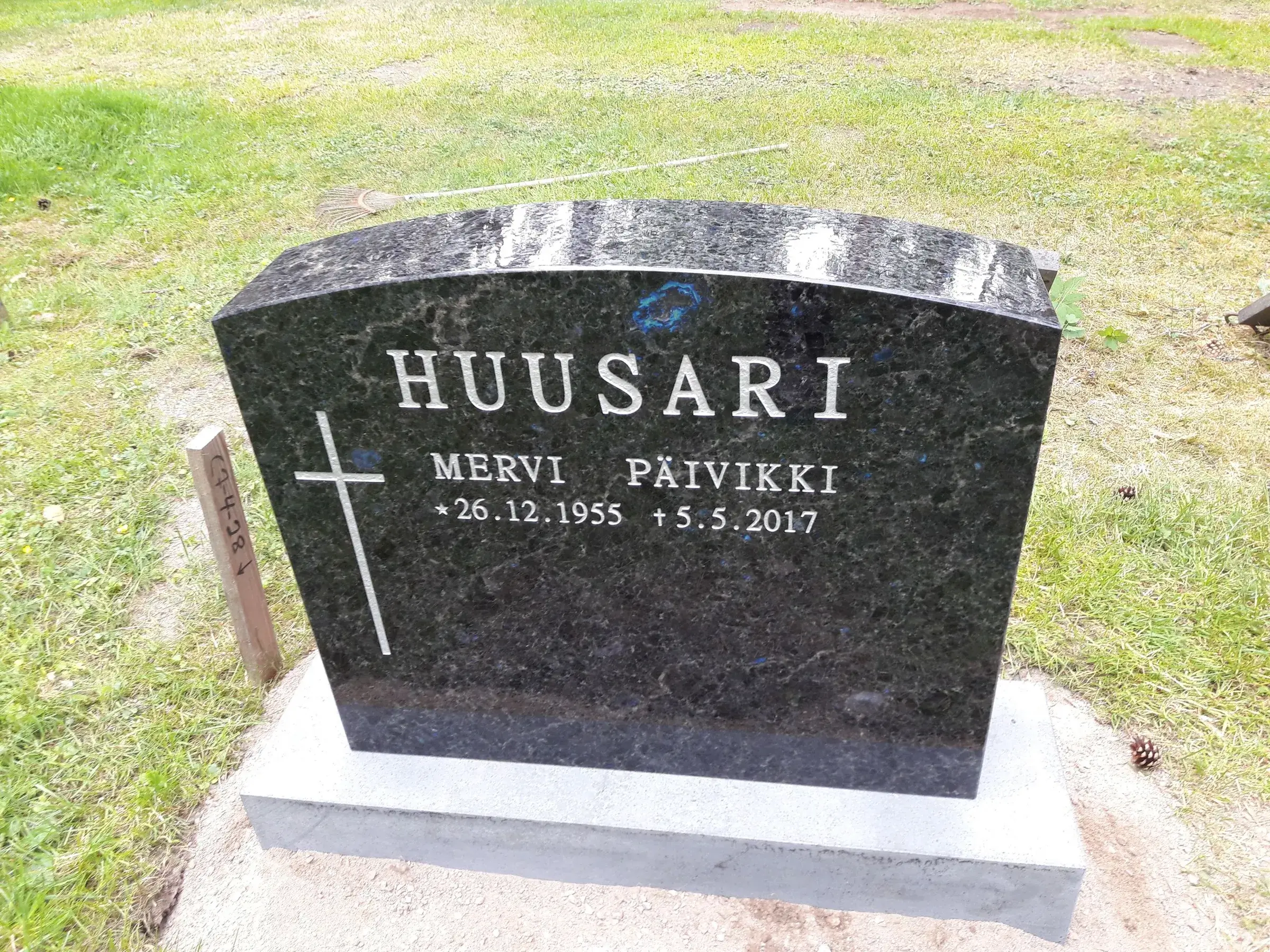 Tumma, kaareva graniittinen hautakivi seisoo vihreällä nurmikolla. Kiveen on kaiverrettu vaalealla tekstillä sukunimi 'HUUSARI', etunimet 'MERVI PÄIVIKKI' sekä syntymä- ja kuolinpäivät '* 26.12.1955 + 5.5.2017'. Vasemmalla on vaalea risti. Hautakivi on vaaleanharmaan jalustan päällä. Taustalla nurmikolla näkyy harava ja oikealla puolella käpy. Vasemmalla on puinen merkintäkeppi, jossa lukee 'C2-4-28'. Kuva on otettu kirkkaassa päivänvalossa.
