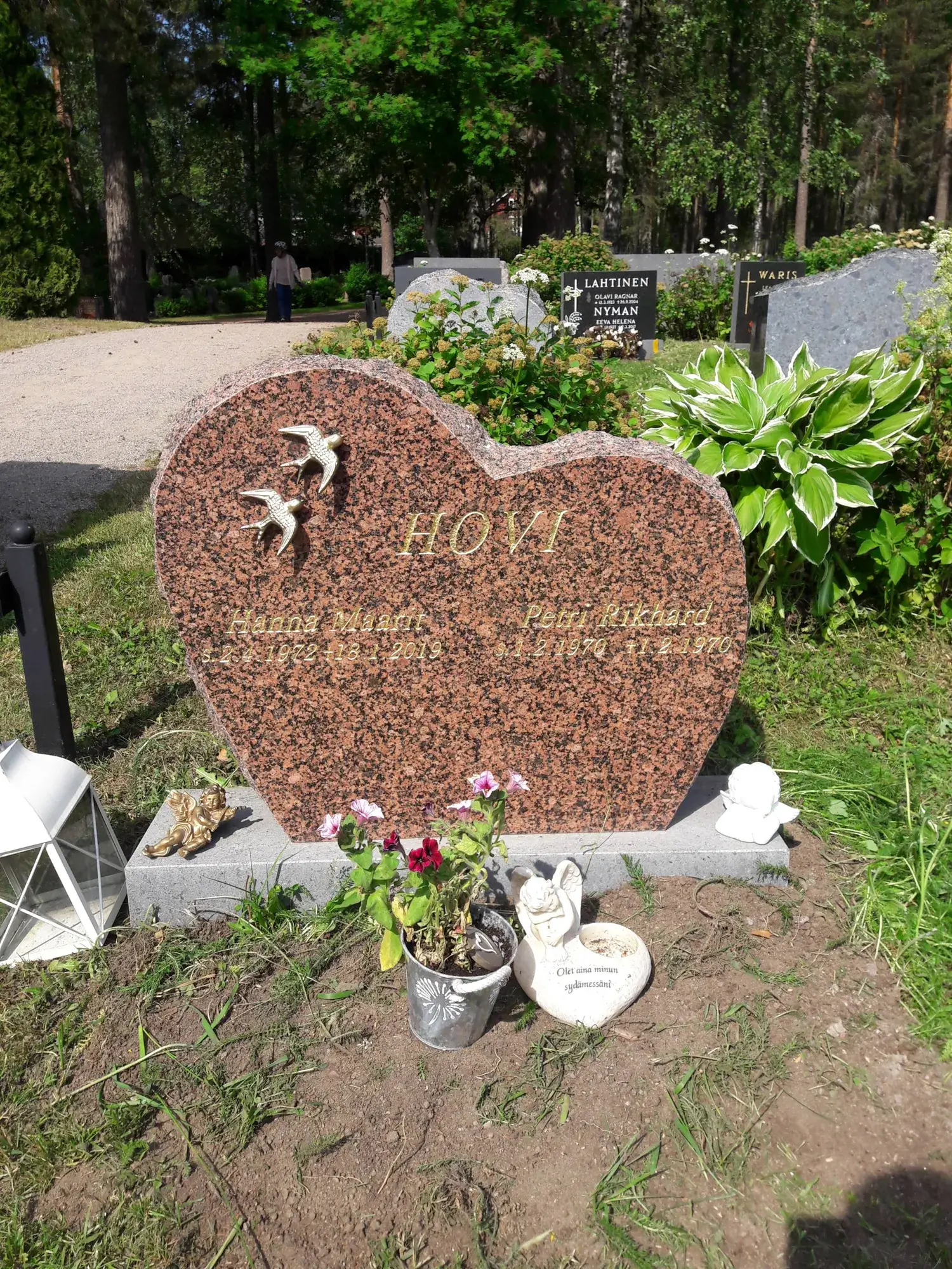 Aurinkoisella hautausmaalla lähikuva sydämenmuotoisesta punaruskeasta graniittisesta hautakivestä. Kivessä on kultainen teksti "HOVI", "Hanna Maarit s. 2.4.1972 + 18.1.2019" ja "Petri Rikhard s. 1.2.1970 + 1.2.1970" sekä kaksi kultaista lintuhahmoa. Hautakiven edustalla on valkoinen lyhty, kultainen ja valkoinen enkelihahmo sekä sydämenmuotoinen kivi, jossa lukee "Olet aina minun sydämessäni". Taustalla näkyy sorapolku, vihreää kasvillisuutta, puita ja muita hautakiviä, ja kaukana kävelee henkilö.
