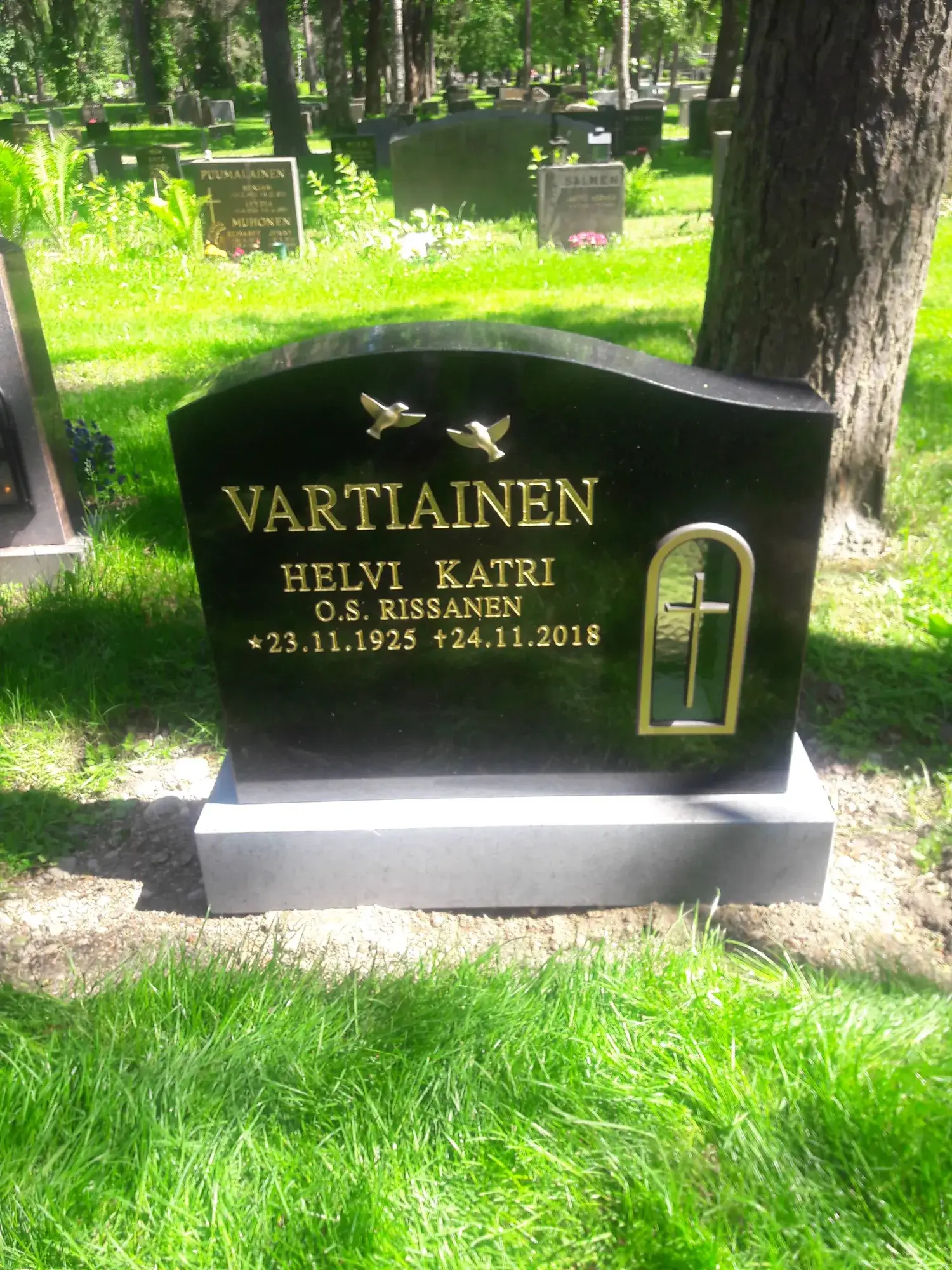 Musta hautakivi kultaisella tekstillä ja symboleilla. Kiven yläosassa on kaksi lentävää kultaista lintua. Teksti lukee 'VARTIAINEN', 'HELVI KATRI', 'O.S. RISSANEN' sekä syntymä- ja kuolinaika '*23.11.1925 +24.11.2018'. Oikealla puolella on kultainen kaari-ikkunan muotoinen kehys, jossa on vihreä tausta ja kultainen risti. Hautakivi seisoo vaaleanharmaalla jalustalla. Kuvan etualalla ja taustalla on vihreää nurmikkoa, ja taustalla näkyy muita hautakiviä ja puita aurinkoisessa hautausmaaympäristössä.