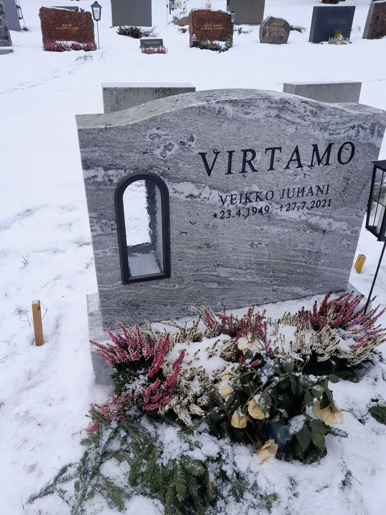 Harmaa graniittinen hautakivi seisoo lumisella hautausmaalla. Kivessä on kaiverrettuna nimi VIRTAMO ja vainajan tiedot: VEIKKO JUHANI *23.4.1949 †27.7.2021. Hautakiven vasemmalla puolella on kaareva, syvennetty aukko, jossa on heijastava lasipinta. Kiven juurella on vaaleanpunaisia ja valkoisia kanervia, vihreitä havunoksia ja muutamia lumen peittämiä vaaleankeltaisia ruusuja. Maa on kauttaaltaan lumen peitossa, ja taustalla näkyy useita muita hautakiviä. Kuvan valaistus on tasainen ja pehmeä.